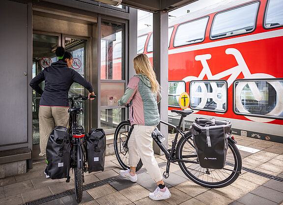 Zwei Frauen schieben zwei Fahrräder in einen Aufzug am Bahnhof. Im Hinterung ein Regionalzug mit Fahrradsymbol.